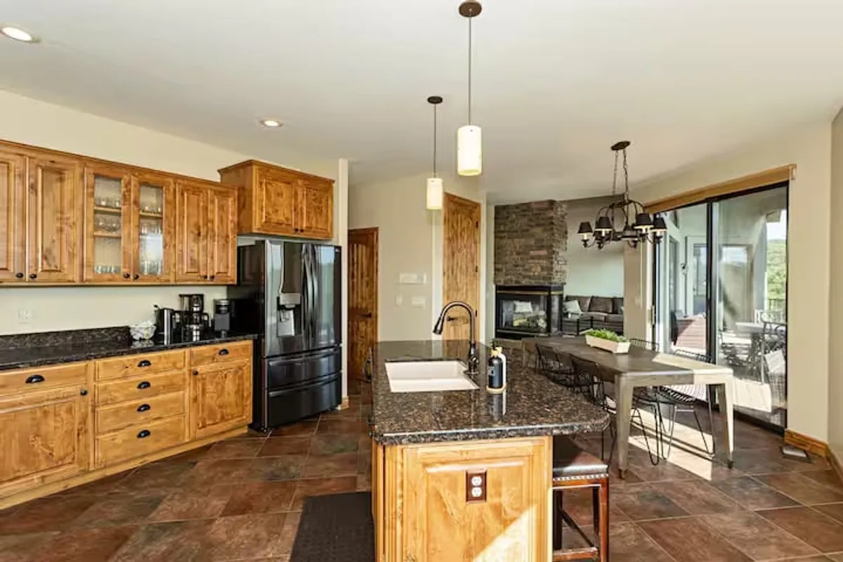 This image captures a kitchen island with stainless steel appliances, a stone fireplace, and a dining area. The open plan allows for easy flow between cooking, dining, and lounging. The scene is warm and ideal for group activities and relaxed family meals.