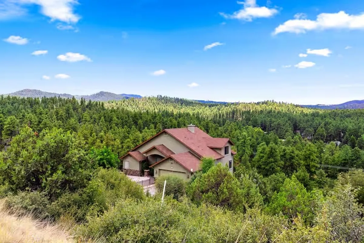 An inviting exterior view of the house, nestled among tall trees with a striking mountain backdrop. The setting exudes tranquility and access to nature, suggesting immediate proximity to trails and outdoor exploration.