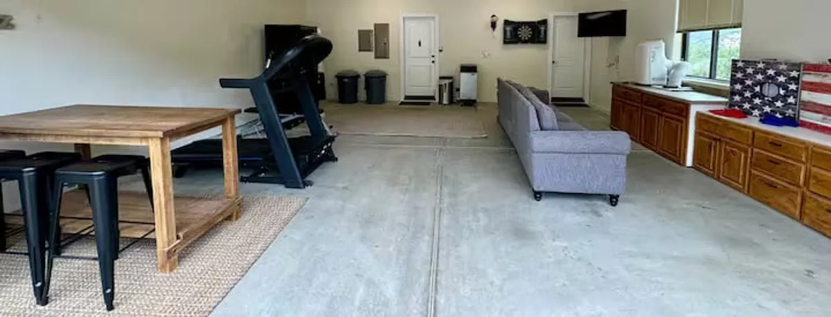 A recreation room featuring a wooden dining table, treadmill, grey couch, American flag decor, and storage cabinets. The layout is casual and functional, supporting multiple activities such as games, exercise, and TV viewing.