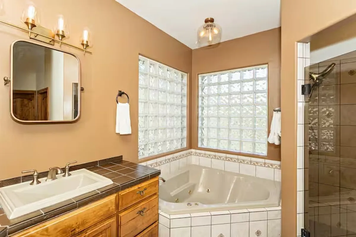A tranquil bathroom scene with a jacuzzi tub, glass block windows allowing in natural light, wooden cabinetry, and a well-lit vanity mirror. The inviting space is designed for indulgent relaxation and self-care.