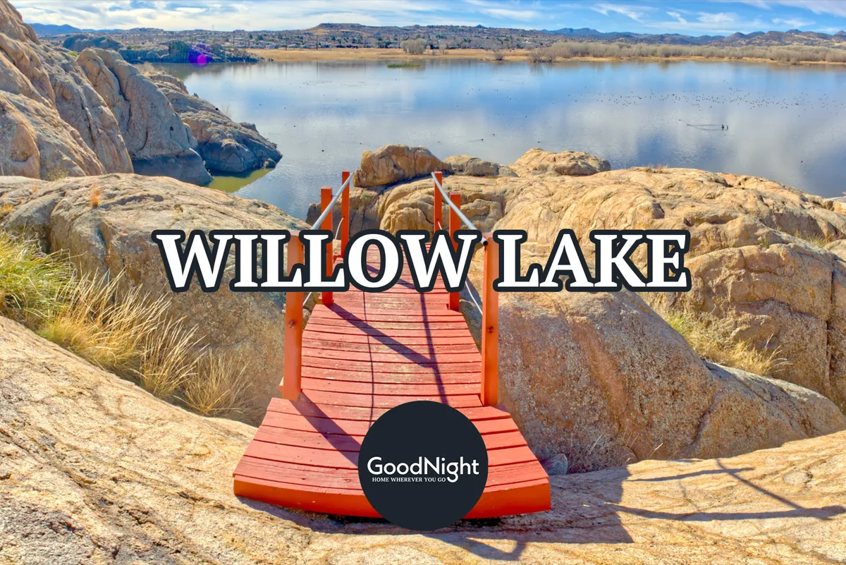 A tranquil scene featuring a red wooden bridge spanning a calm lake, surrounded by rugged rock formations and distant hills. The scenery is alive with possibilities for hiking, fishing, and observing wildlife, especially during a beautiful sunset.