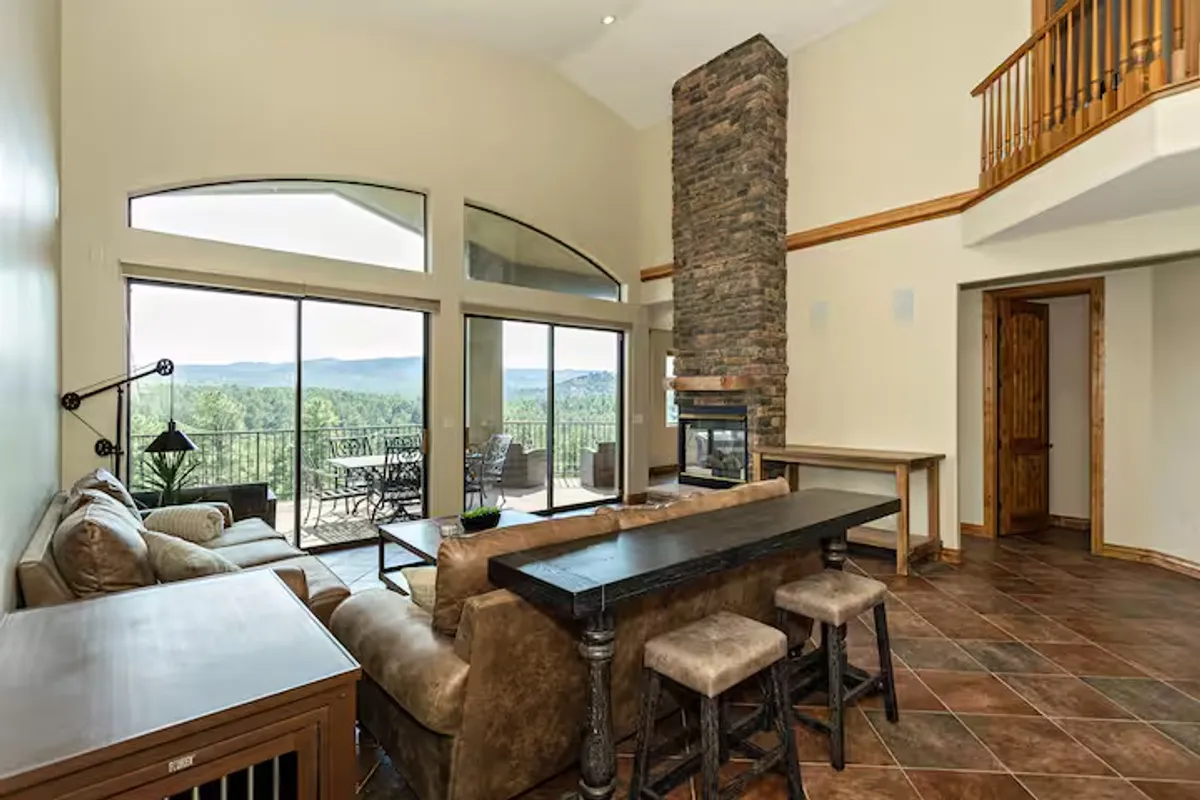 This image portrays a spacious, open-concept living area with large windows overlooking the mountains, a stone fireplace, a comfortable sofa, and a dining table with bar stools. The setup visually connects lounging, dining, and kitchen areas within one inviting space, fostering togetherness and interaction.