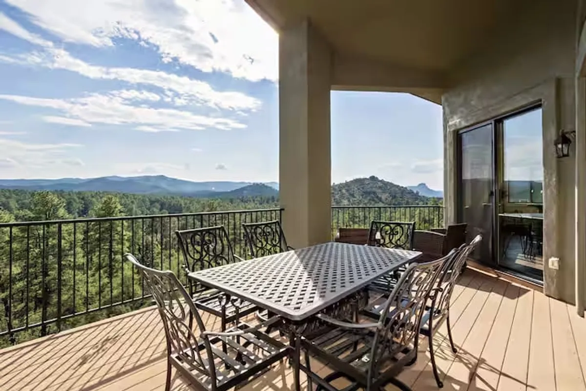 An outdoor dining table is set on a balcony with sturdy railings, overlooking distant mountain views. The table is arranged for relaxed, social meals, and the atmosphere is tranquil and inviting, with natural light and fresh air enhancing the setting.
